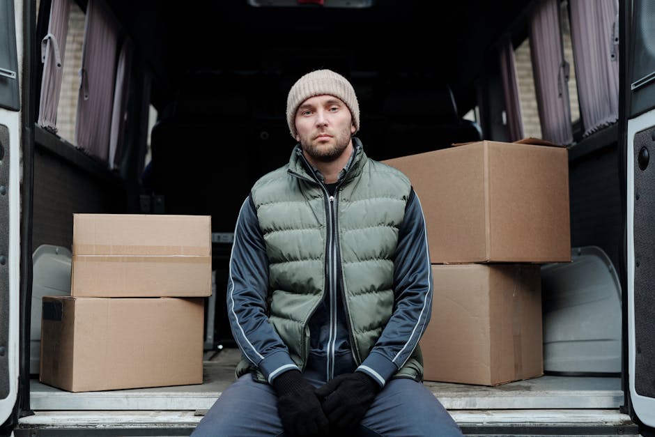 A man wearing a dark jacket and cap is seen loading or unloading cardboard boxes from a white moving van parked outside a modern building with large glass windows. The open van door reveals additional