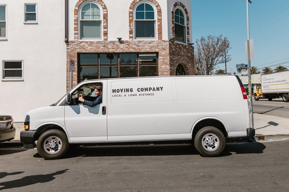 A white moving van parked on a city street in front of a brick building with large windows, clearly marked with 'MOVING COMPANY LOCAL & LONG DISTANCE' on its side. The driver, wearing sunglasses and a dark jacket, is seated in the cab and appears to be preparing or checking the vehicle. The van is positioned parallel to the sidewalk, with the pavement visible beneath it. In the background, there are other vehicles, including a yellow truck and a smaller car, along with a tree and street signs indicating urban surroundings. The scene depicts a home relocation process, with the van likely involved in furniture transport and packing and moving activities, and is set during daylight hours with bright, natural lighting. This image relates to professional removals services offered by Man with Van Streatham, featuring the loading process typical of house moves and furniture transportation within an urban environment.
